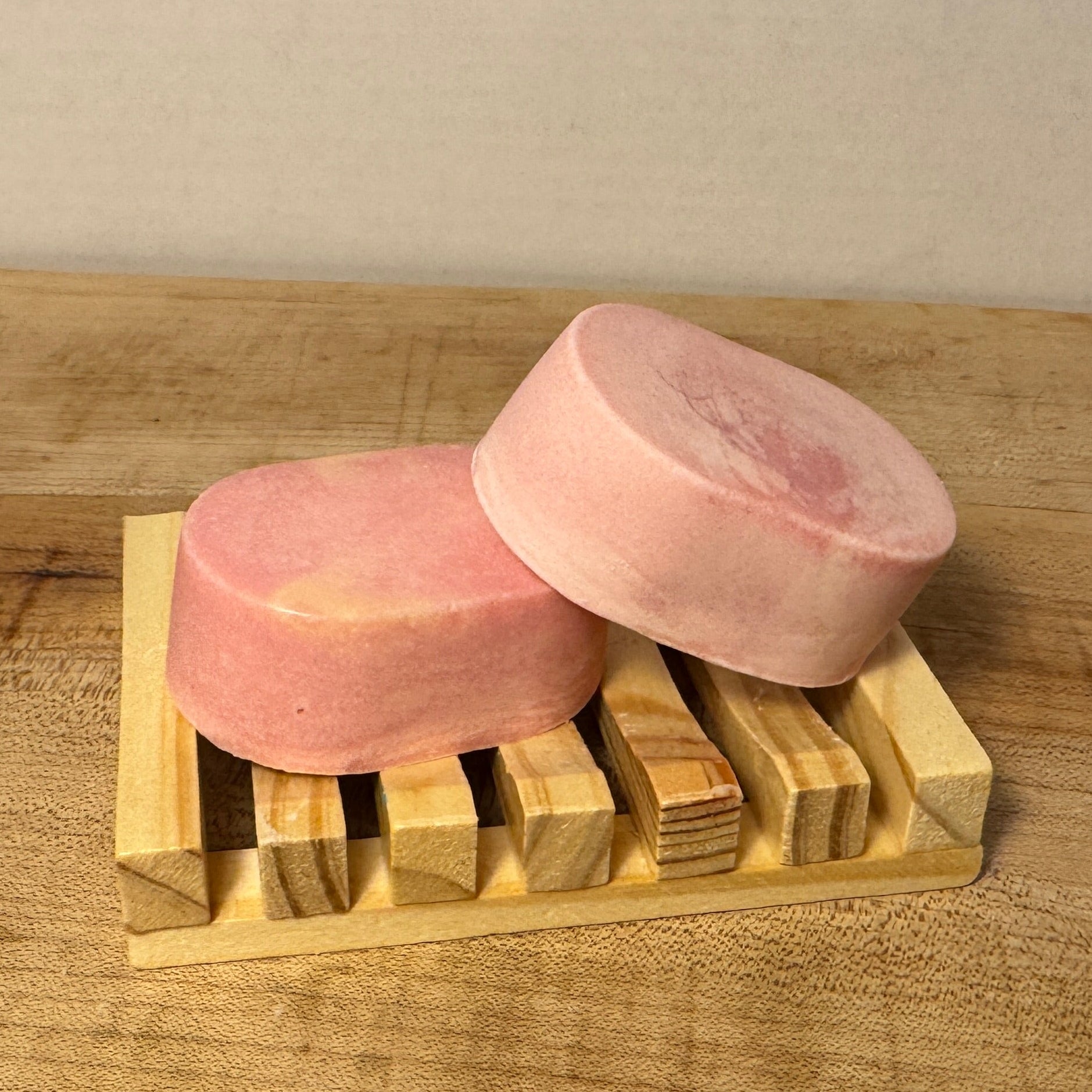 Two pink soap bars on a wooden soap dish against a plain background