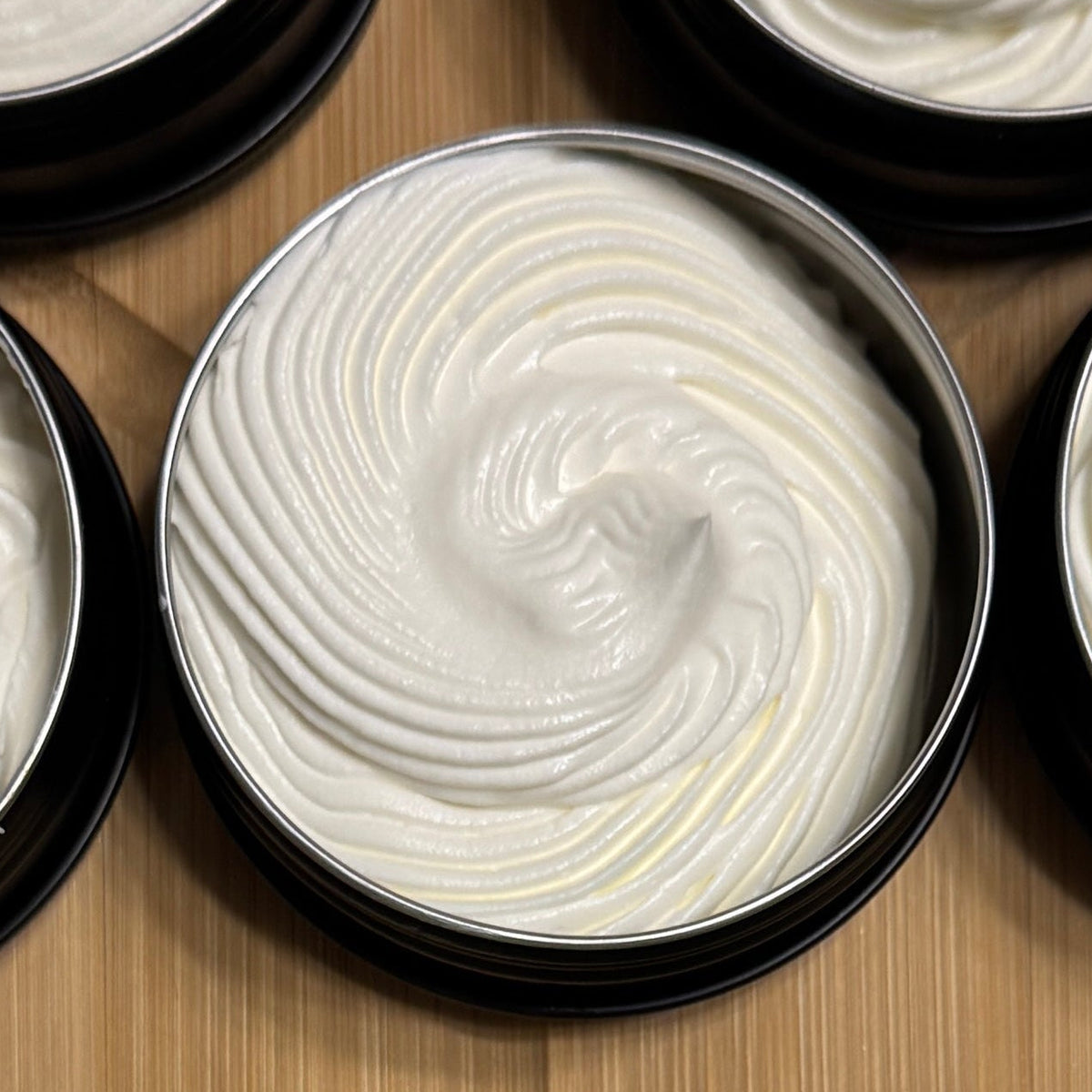 Nine small round metal jars filled with whipped white cream or lotion, arranged on a light wood surface.