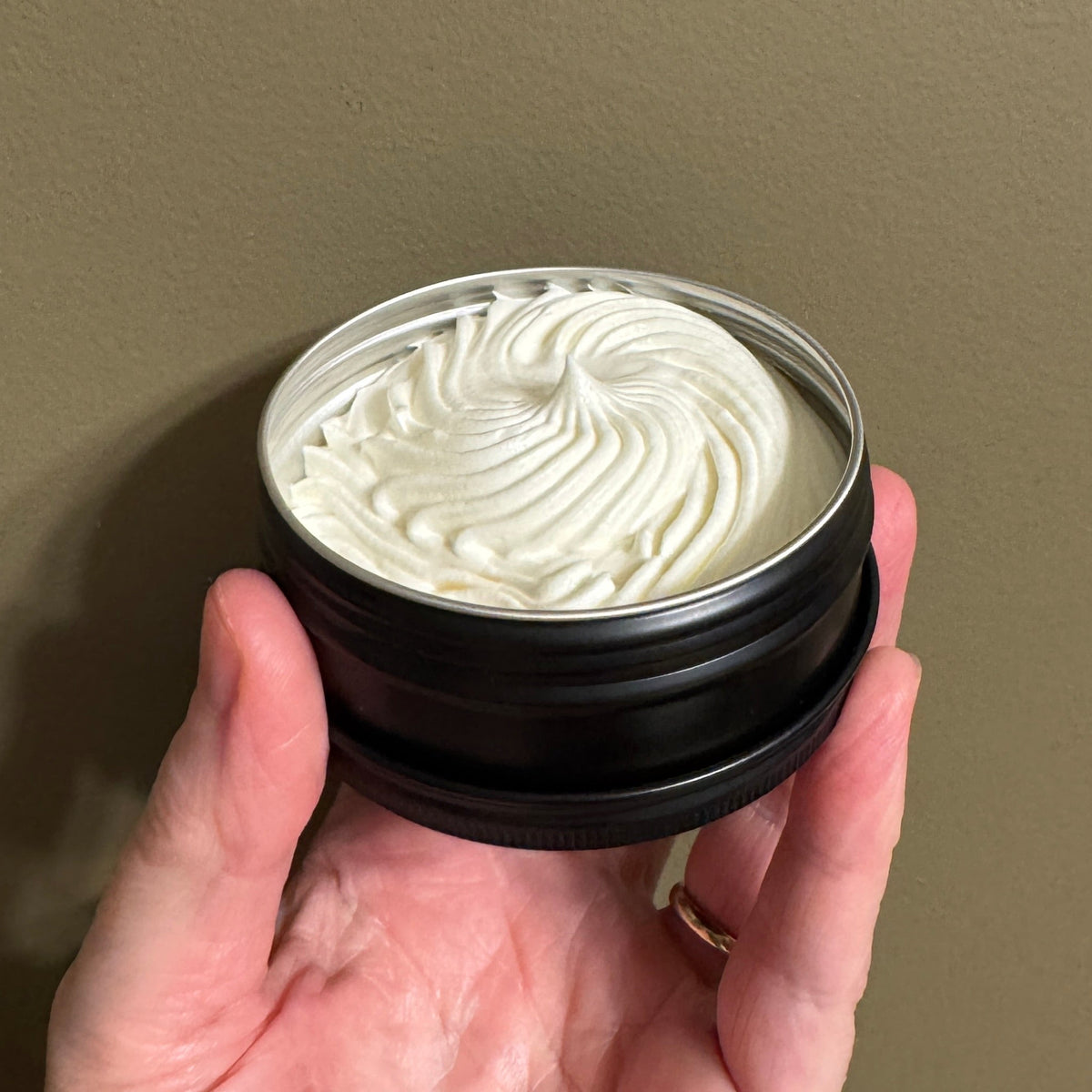 A hand holding an open round black tin showing a whipped off-white cream product against a plain taupe wall.