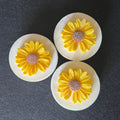 Three round white soaps with yellow daisy flower charms in the center, displayed on a dark gray textured background.