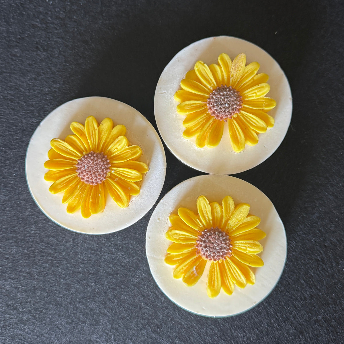 Three round white soaps with yellow daisy flower charms in the center, displayed on a dark gray textured background.