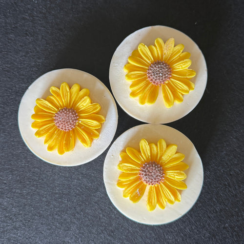 Three round white soaps with yellow daisy flower charms in the center, displayed on a dark gray textured background.